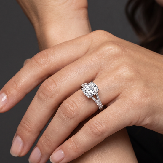 A close-up of a woman's hand resting gracefully against her arm, showcasing a large, elongated cushion-cut diamond engagement ring with a diamond-encrusted split-shank band against a dark gray background.