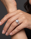 A close-up of a woman's hand resting gracefully against her arm, showcasing a large, elongated cushion-cut diamond engagement ring with a diamond-encrusted split-shank band against a dark gray background.