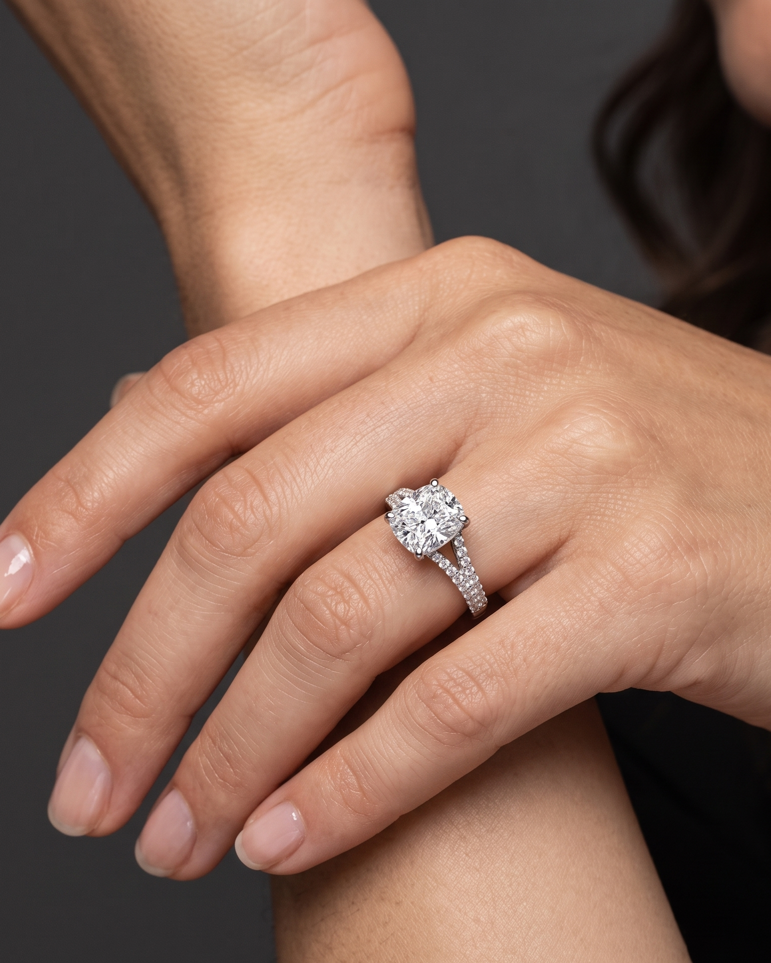 A close-up of a woman's hand resting gracefully against her arm, showcasing a large, elongated cushion-cut diamond engagement ring with a diamond-encrusted split-shank band against a dark gray background.