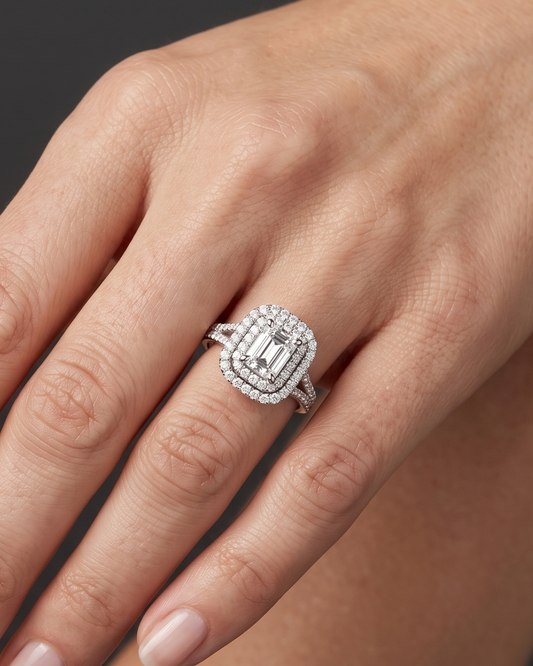 A close-up of a woman's elegantly posed hand wearing a luxurious white gold engagement ring with an emerald-cut center diamond and a double halo, set against a dark charcoal gray background.
