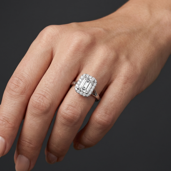A close-up shot of a woman's hand resting gracefully against a smooth, dark charcoal gray background. On her ring finger, she wears an elegant silver-toned engagement ring featuring a large emerald-cut center diamond surrounded by a halo of smaller round diamonds on a split-shank band. Soft studio lighting highlights the ring's brilliant sparkle and the natural details of her skin.