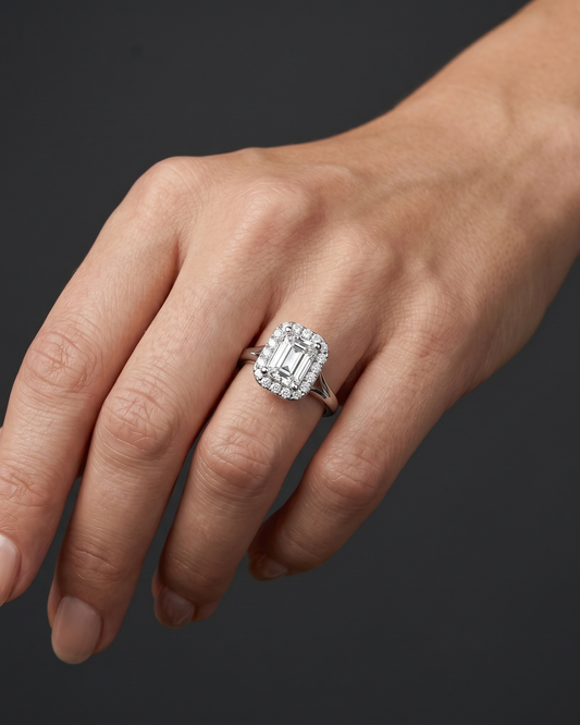 A close-up shot of a woman's hand resting gracefully against a smooth, dark charcoal gray background. On her ring finger, she wears an elegant silver-toned engagement ring featuring a large emerald-cut center diamond surrounded by a halo of smaller round diamonds on a split-shank band. Soft studio lighting highlights the ring's brilliant sparkle and the natural details of her skin.