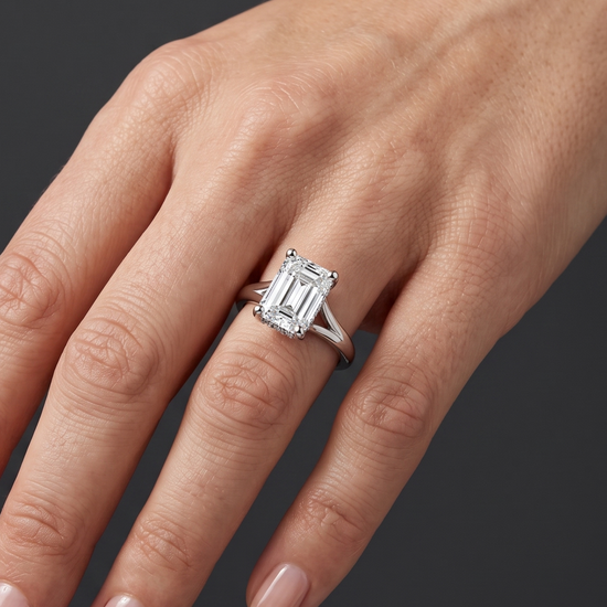 Close-up of a woman's hand wearing an emerald-cut diamond engagement ring with a split-shank band against a dark charcoal gray background.
