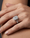 Elegant close-up of a woman's manicured hand with nude polish, resting gently on an arm. On her ring finger, a brilliant emerald-cut diamond ring with a halo setting is prominently displayed.