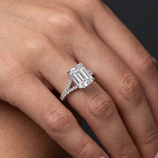 A close-up of a woman's hand wearing a large emerald-cut diamond engagement ring with a diamond-encrusted split-shank band, set against a dark gray background.