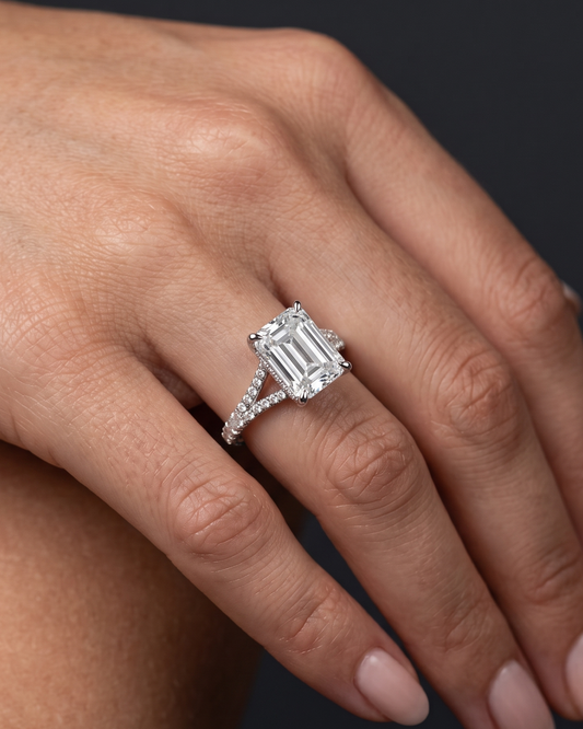 A close-up of a woman's hand wearing a large emerald-cut diamond engagement ring with a diamond-encrusted split-shank band, set against a dark gray background.