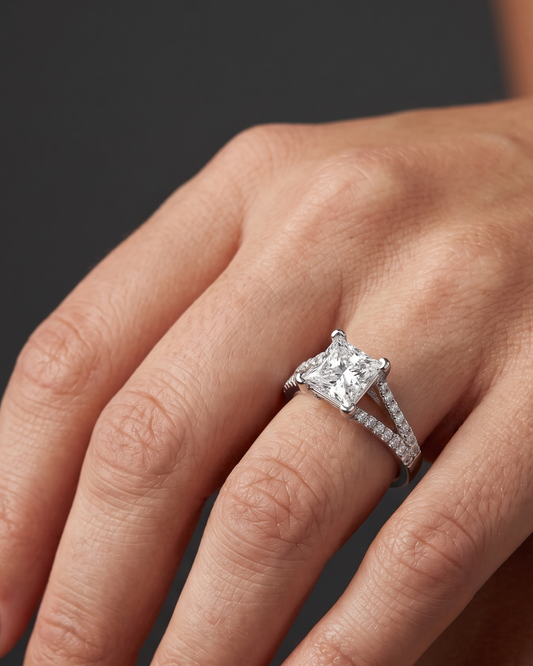 A close-up editorial photograph of a woman's elegantly posed hand wearing a white gold engagement ring. The ring features a prominent princess-cut center diamond set on a diamond-encrusted, split-shank band. The hand rests softly against a smooth, dark charcoal gray background under diffused studio lighting.