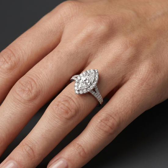 A close-up of a woman's elegantly posed hand resting against a smooth, dark charcoal gray background, showcasing a luxury diamond ring. The ring features a large marquise-cut center diamond surrounded by a halo of smaller round diamonds, set on a split-shank band paved with additional diamonds in a polished white metal.
