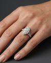 A close-up of a woman's elegantly posed hand resting against a smooth, dark charcoal gray background, showcasing a luxury diamond ring. The ring features a large marquise-cut center diamond surrounded by a halo of smaller round diamonds, set on a split-shank band paved with additional diamonds in a polished white metal.