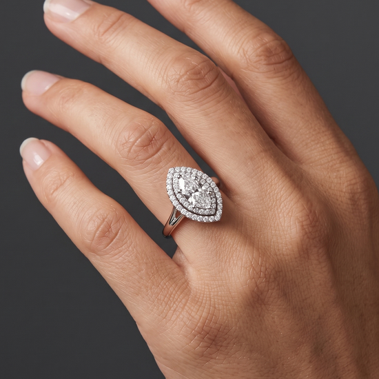 A close-up of a woman's elegantly posed hand against a smooth, dark charcoal gray background, wearing a brilliant engagement ring on her ring finger. The ring features a marquise-cut center diamond surrounded by a double halo of smaller diamonds, set on a polished white gold or platinum band.