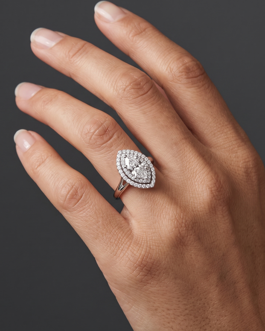 A close-up of a woman's elegantly posed hand against a smooth, dark charcoal gray background, wearing a brilliant engagement ring on her ring finger. The ring features a marquise-cut center diamond surrounded by a double halo of smaller diamonds, set on a polished white gold or platinum band.
