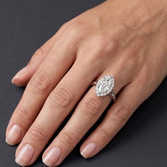 A close-up of an elegant woman's hand resting on a dark charcoal gray surface. She is wearing a brilliant marquise-cut diamond engagement ring with a diamond halo on her ring finger. Soft, diffused lighting highlights the sparkling gemstone, the polished silver band, and the natural texture of her skin.
