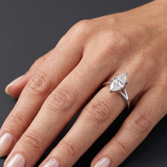A close-up of a woman's hand resting on a dark gray background, wearing a white metal engagement ring with a marquise-cut diamond.