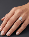A close-up of a woman's hand resting on a dark gray background, wearing a white metal engagement ring with a marquise-cut diamond.