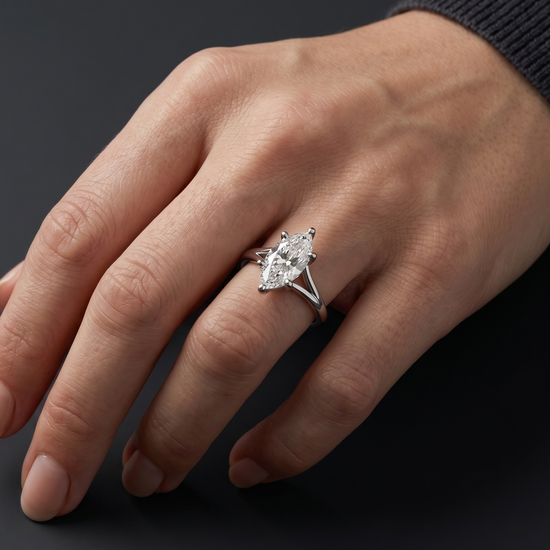 Close-up of a woman's elegantly posed hand wearing a stunning 3-carat marquise-cut diamond solitaire ring with a split shank setting, against a dark charcoal gray background.