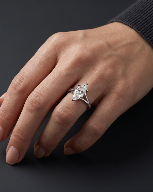 Close-up of a woman's elegantly posed hand wearing a stunning 3-carat marquise-cut diamond solitaire ring with a split shank setting, against a dark charcoal gray background.