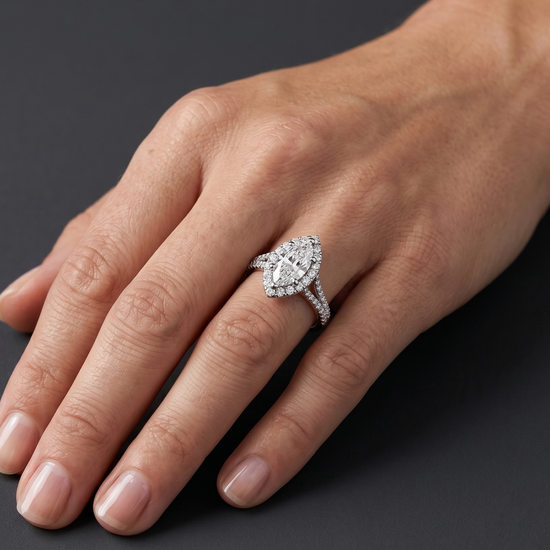 A close-up, luxury editorial shot of a woman's hand resting elegantly against a smooth, dark charcoal gray background. On her ring finger, she is wearing a sparkling diamond ring featuring a large marquise-cut center stone surrounded by a diamond halo, set on a diamond-paved split-shank band. The soft, diffused studio lighting highlights the brilliance of the diamonds, the polished white metal, and the natural texture of her skin.