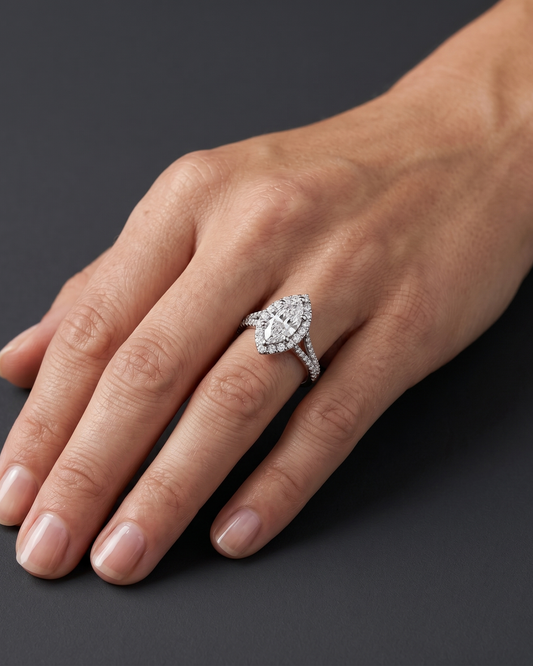 A close-up, luxury editorial shot of a woman's hand resting elegantly against a smooth, dark charcoal gray background. On her ring finger, she is wearing a sparkling diamond ring featuring a large marquise-cut center stone surrounded by a diamond halo, set on a diamond-paved split-shank band. The soft, diffused studio lighting highlights the brilliance of the diamonds, the polished white metal, and the natural texture of her skin.
