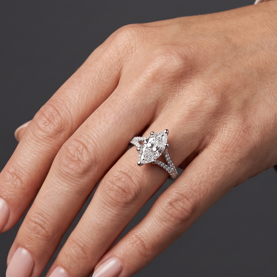 A close-up of a woman's hand elegantly posed against a dark charcoal gray background, wearing a white-gold engagement ring with a large marquise-cut diamond and a pavé split-shank band.