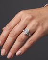 A close-up of a woman's hand elegantly posed against a dark charcoal gray background, wearing a white-gold engagement ring with a large marquise-cut diamond and a pavé split-shank band.