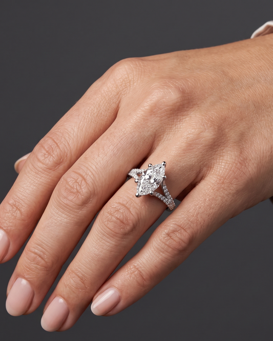 A close-up of a woman's hand elegantly posed against a dark charcoal gray background, wearing a white-gold engagement ring with a large marquise-cut diamond and a pavé split-shank band.