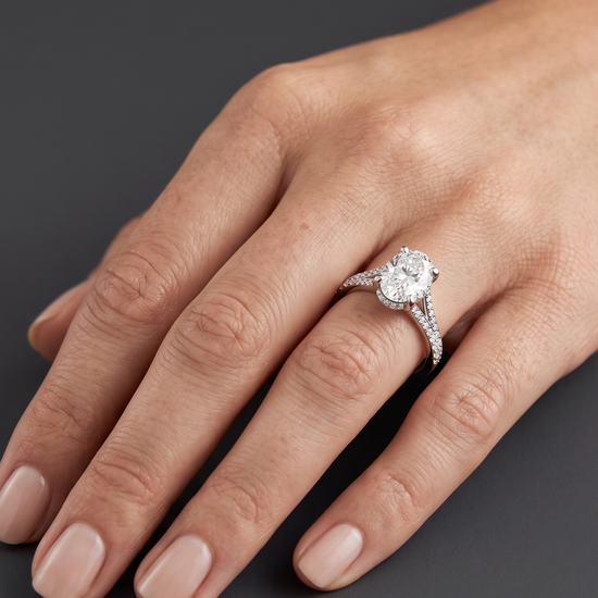 Close-up of a woman's hand resting elegantly against a smooth, dark charcoal gray background, showcasing a brilliant engagement ring. The ring features a large oval-cut center diamond set in white gold or platinum, detailed with a diamond-encrusted split-shank band and a hidden halo.