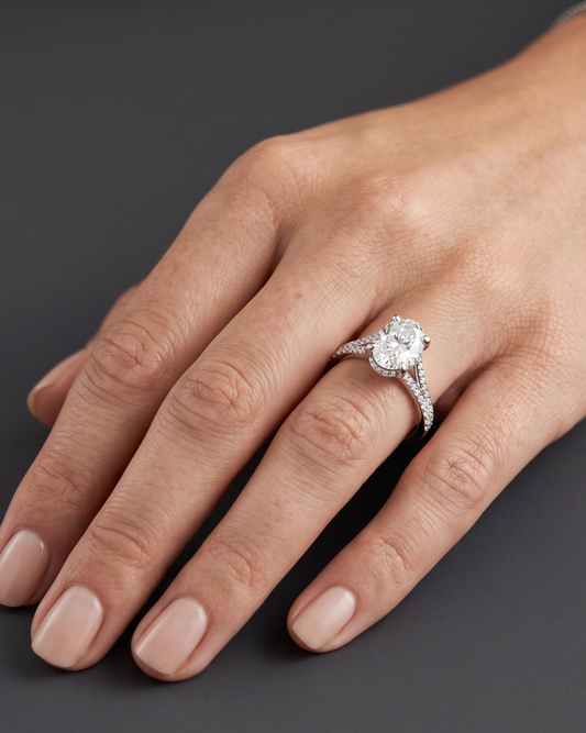 Close-up of a woman's hand resting elegantly against a smooth, dark charcoal gray background, showcasing a brilliant engagement ring. The ring features a large oval-cut center diamond set in white gold or platinum, detailed with a diamond-encrusted split-shank band and a hidden halo.