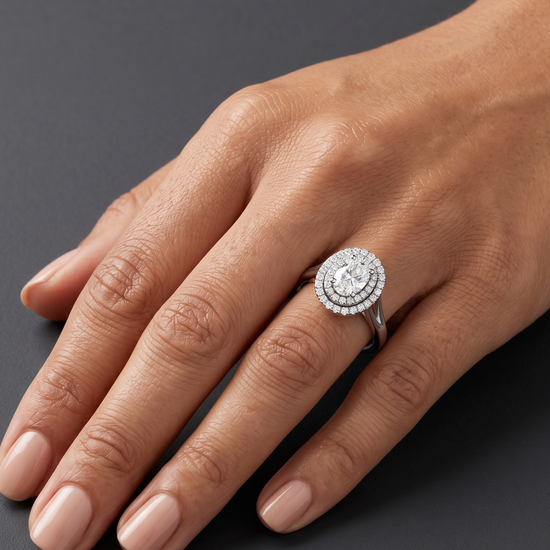 An editorial close-up of a woman's hand resting on a smooth dark gray surface to showcase a luxury engagement ring. The ring features a large oval center diamond surrounded by a double halo of smaller diamonds on a silver-toned split-shank band.