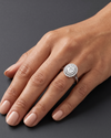 An editorial close-up of a woman's hand resting on a smooth dark gray surface to showcase a luxury engagement ring. The ring features a large oval center diamond surrounded by a double halo of smaller diamonds on a silver-toned split-shank band.