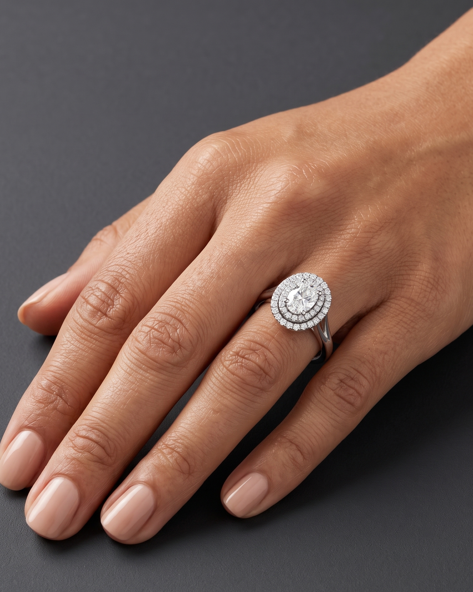 An editorial close-up of a woman's hand resting on a smooth dark gray surface to showcase a luxury engagement ring. The ring features a large oval center diamond surrounded by a double halo of smaller diamonds on a silver-toned split-shank band.