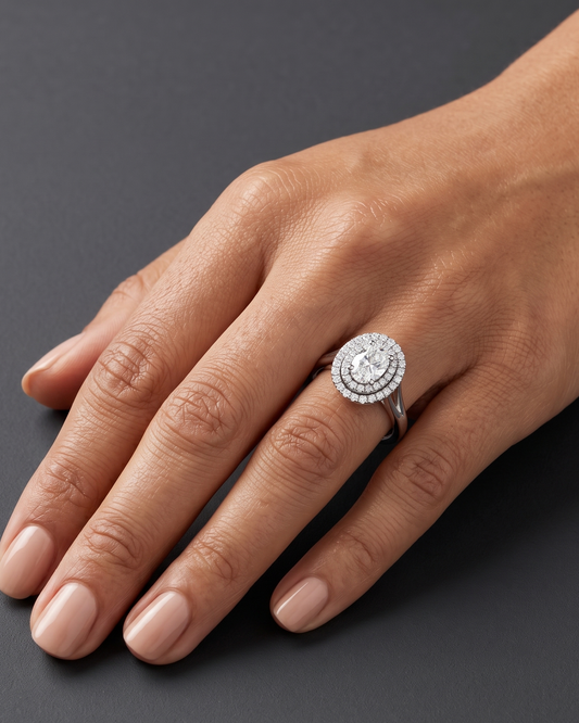 An editorial close-up of a woman's hand resting on a smooth dark gray surface to showcase a luxury engagement ring. The ring features a large oval center diamond surrounded by a double halo of smaller diamonds on a silver-toned split-shank band.