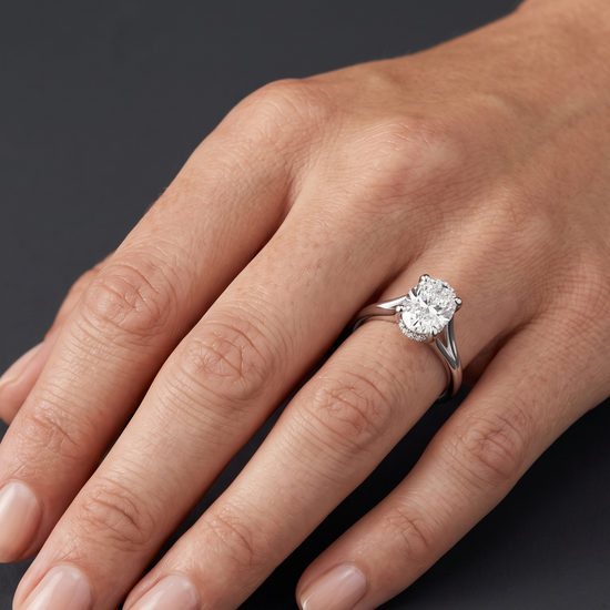 A close-up of a woman's hand resting gracefully against a dark charcoal gray background, wearing a silver-tone engagement ring with an oval-cut diamond and a split-shank band.
