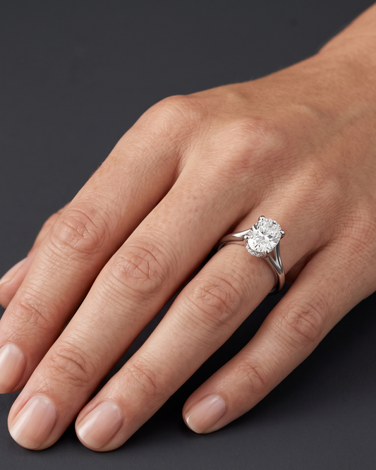 A close-up of a woman's hand resting gracefully against a dark charcoal gray background, wearing a silver-tone engagement ring with an oval-cut diamond and a split-shank band.