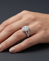 A close-up editorial shot of a woman's elegantly posed hand resting on a smooth, dark charcoal gray surface. She is wearing a luxury white-metal engagement ring on her ring finger, featuring a large oval-cut center diamond surrounded by a diamond halo, with a diamond-paved split-shank band. Soft studio lighting highlights the brilliant sparkle of the gemstone and the natural texture of her skin. Her nails are manicured in a soft, neutral shade.