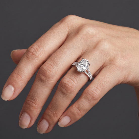A close-up of a woman's hand wearing a silver engagement ring with an oval-cut center diamond and a diamond-encrusted split band, set against a dark gray background.