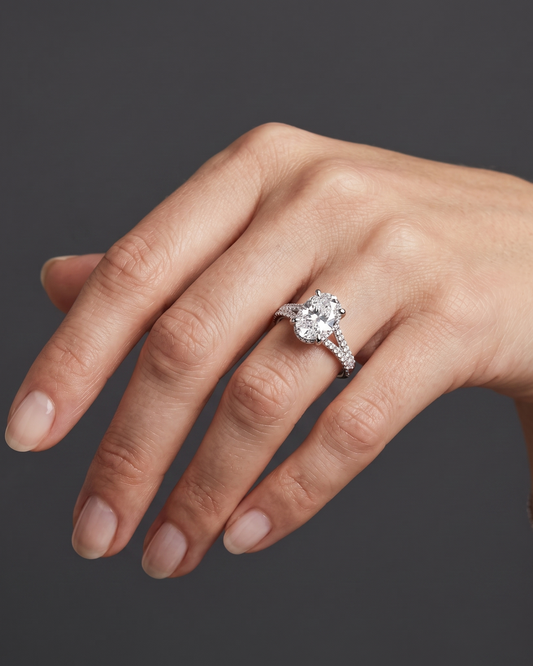 A close-up of a woman's hand wearing a silver engagement ring with an oval-cut center diamond and a diamond-encrusted split band, set against a dark gray background.
