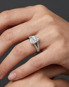 A close-up, hyper-realistic editorial photograph of a woman's hands gracefully posed against a smooth, dark charcoal gray background. On her ring finger, she is wearing a luxury silver-toned engagement ring featuring a large oval-cut center diamond and a pave diamond-encrusted split-shank band. The soft, diffused studio lighting highlights the intricate details and sparkle of the ring, as well as the natural texture and skin compression of her hands.