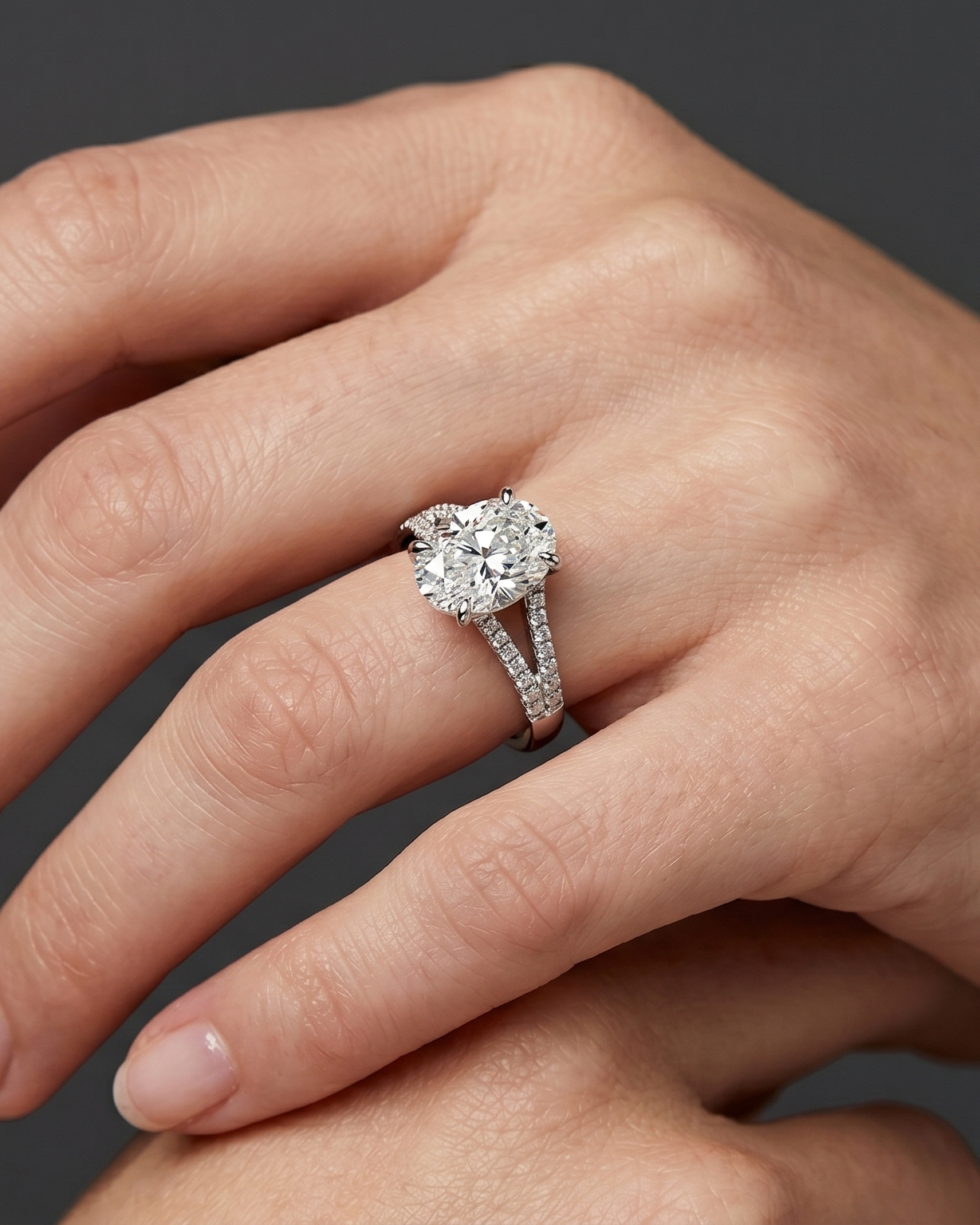 A close-up, hyper-realistic editorial photograph of a woman's hands gracefully posed against a smooth, dark charcoal gray background. On her ring finger, she is wearing a luxury silver-toned engagement ring featuring a large oval-cut center diamond and a pave diamond-encrusted split-shank band. The soft, diffused studio lighting highlights the intricate details and sparkle of the ring, as well as the natural texture and skin compression of her hands.