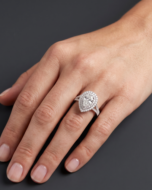 A close-up editorial photograph of a woman's hand resting elegantly on a smooth, dark charcoal gray surface. She is wearing a luxurious white gold or platinum diamond ring on her ring finger. The ring features a brilliant pear-shaped center diamond surrounded by a double halo of smaller diamonds, with the point of the pear shape facing downward toward her wrist. The band is also encrusted with diamonds.