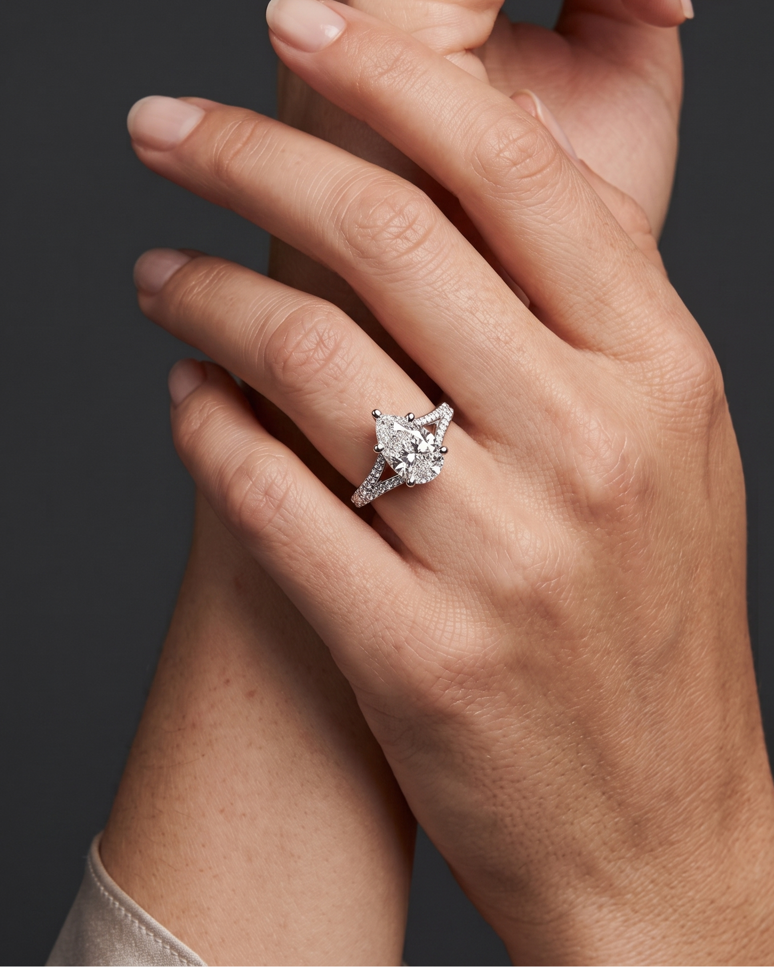 Editorial close-up of a woman's elegantly crossed hands, highlighting a brilliant pear-shaped diamond ring with a diamond-encrusted split-shank band. The photo features soft, realistic lighting and natural skin texture against a smooth, dark gray background.