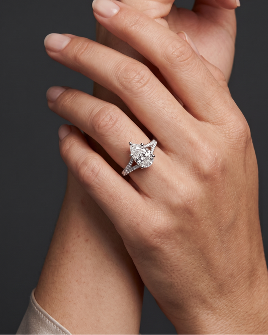 Editorial close-up of a woman's elegantly crossed hands, highlighting a brilliant pear-shaped diamond ring with a diamond-encrusted split-shank band. The photo features soft, realistic lighting and natural skin texture against a smooth, dark gray background.