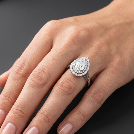 Close-up of a woman's hand resting gracefully against a dark charcoal gray background, wearing a large pear-shaped diamond ring with a double diamond halo setting.
