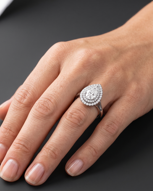 Close-up of a woman's hand resting gracefully against a dark charcoal gray background, wearing a large pear-shaped diamond ring with a double diamond halo setting.
