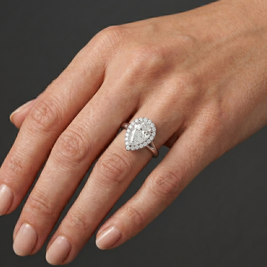 A close-up, editorial-style photograph of a woman's elegantly posed hand resting against a seamless dark charcoal gray background. On her ring finger, she wears a white metal engagement ring featuring a large pear-shaped center diamond surrounded by a diamond halo on a split-shank band. The point of the pear-shaped diamond is oriented downward, pointing toward her wrist. Soft, diffused studio lighting highlights the realistic texture of her skin and the brilliant, sparkling facets of the gemstone and metal.