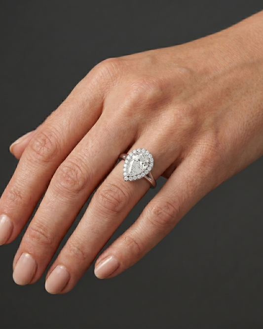 A close-up, editorial-style photograph of a woman's elegantly posed hand resting against a seamless dark charcoal gray background. On her ring finger, she wears a white metal engagement ring featuring a large pear-shaped center diamond surrounded by a diamond halo on a split-shank band. The point of the pear-shaped diamond is oriented downward, pointing toward her wrist. Soft, diffused studio lighting highlights the realistic texture of her skin and the brilliant, sparkling facets of the gemstone and metal.