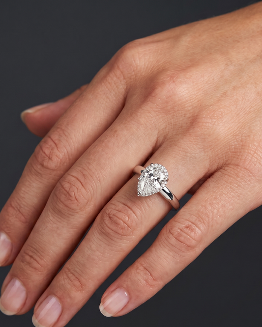 A well-lit, close-up photograph of a manicured left hand, with a medium skin tone, angled slightly against a dark, textured charcoal background. On the ring finger is a white gold engagement ring with a prominent, brilliant pear-shaped diamond, surrounded by a delicate halo of smaller round-cut diamonds. The fingernails are clean and painted with a subtle, clear polish.