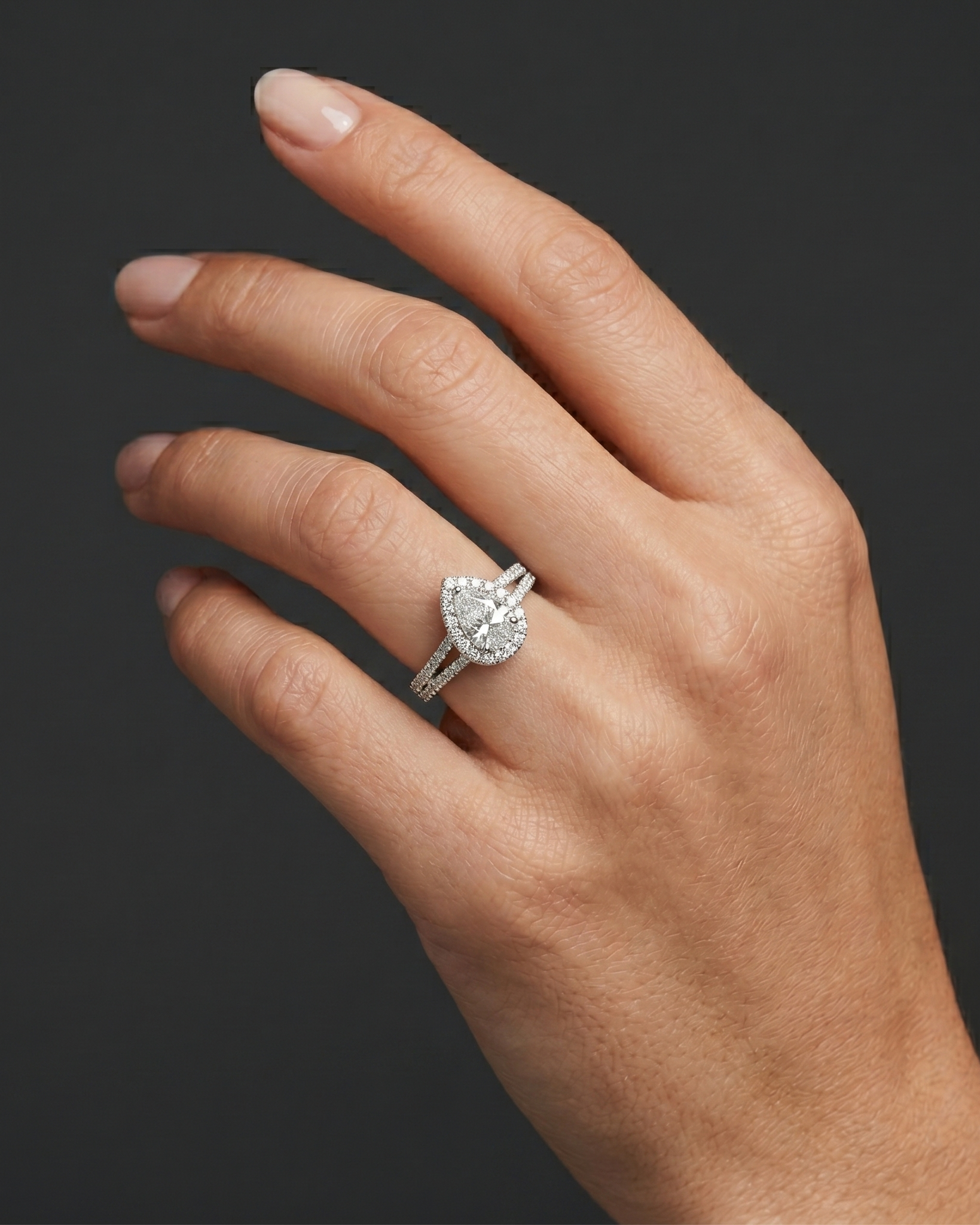 A hyper-realistic close-up of a woman's elegantly posed hand wearing a sparkling, pear-shaped diamond engagement ring. The ring features a diamond halo and a diamond-encrusted split shank, scaled proportionally to sit naturally on her ring finger. The photograph is shot in a luxury editorial style with soft, diffused lighting that highlights both the natural skin texture and the brilliant details of the jewelry, all set against a smooth, dark charcoal gray background.