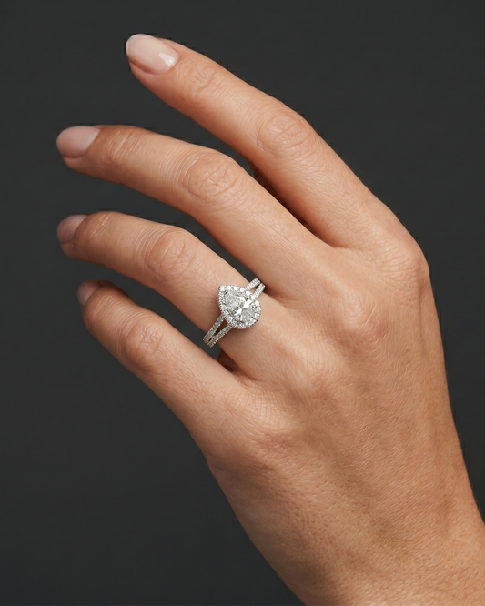 A hyper-realistic close-up of a woman's elegantly posed hand wearing a sparkling, pear-shaped diamond engagement ring. The ring features a diamond halo and a diamond-encrusted split shank, scaled proportionally to sit naturally on her ring finger. The photograph is shot in a luxury editorial style with soft, diffused lighting that highlights both the natural skin texture and the brilliant details of the jewelry, all set against a smooth, dark charcoal gray background.