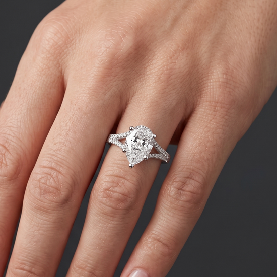 A close-up of a woman's hand wearing a pear-shaped diamond engagement ring with a diamond-encrusted split-shank band. The point of the diamond is facing downward toward her fingertips, set against a dark charcoal gray background.
