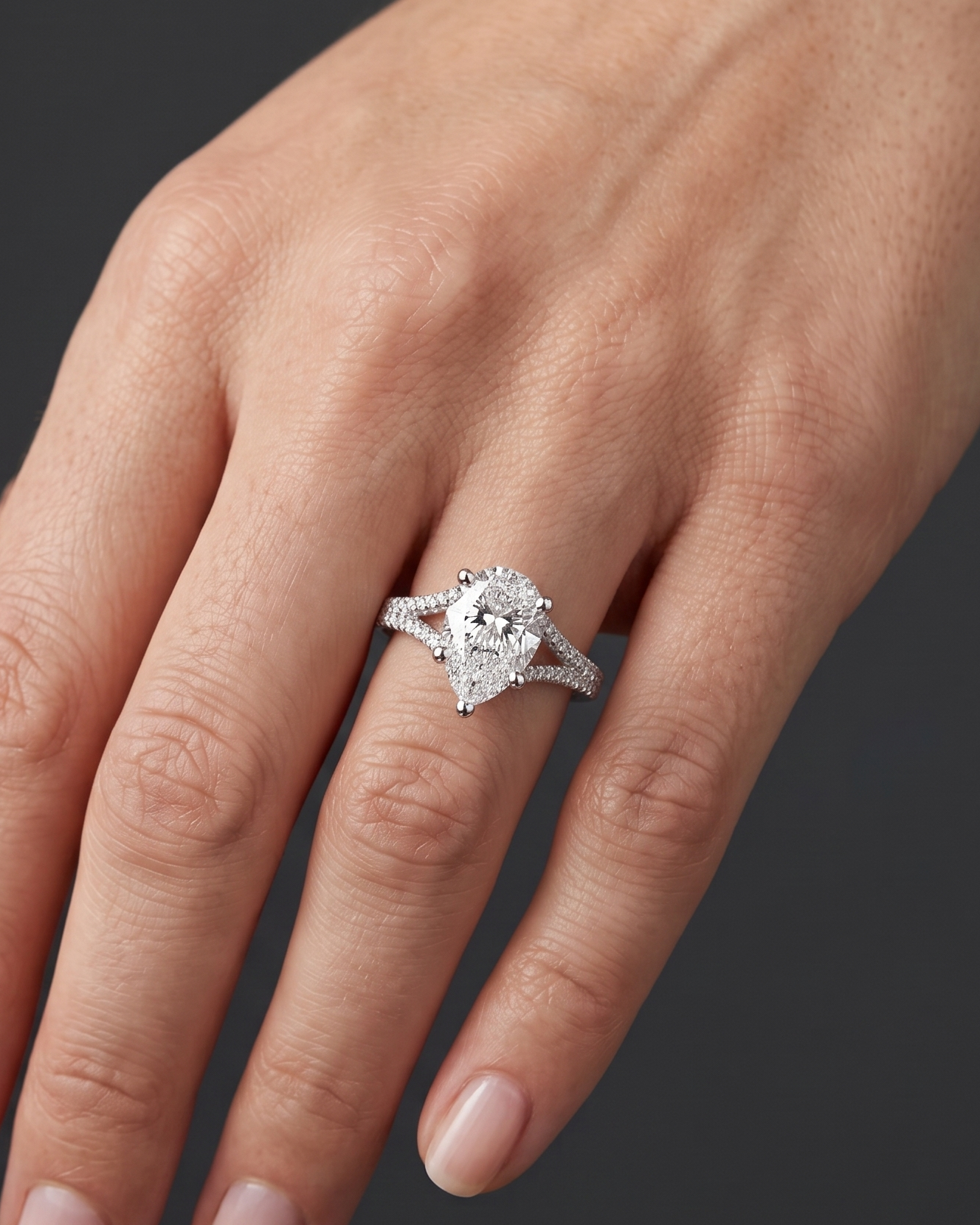 A close-up of a woman's hand wearing a pear-shaped diamond engagement ring with a diamond-encrusted split-shank band. The point of the diamond is facing downward toward her fingertips, set against a dark charcoal gray background.